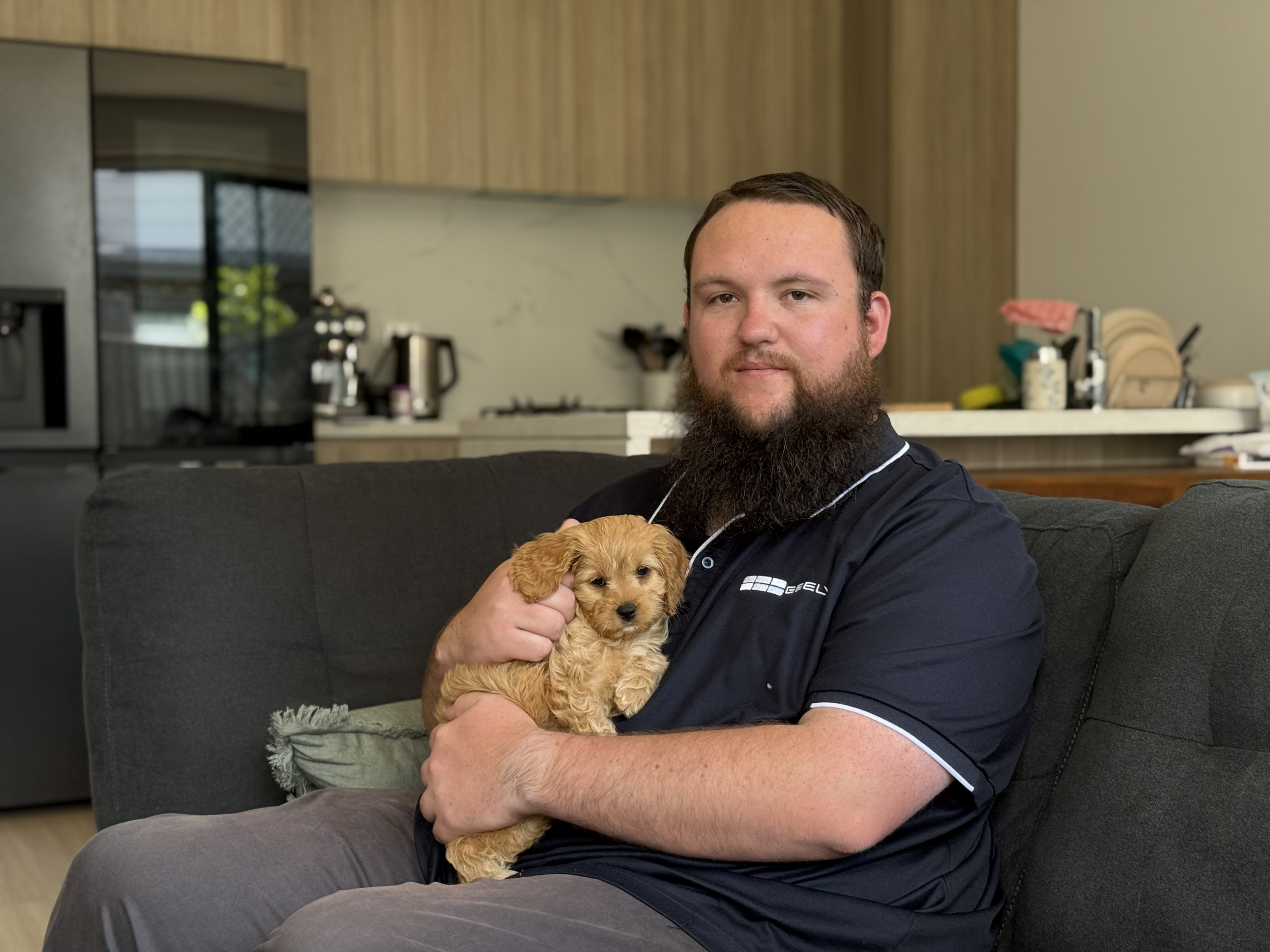 man with beard in polo shirt holds small puppy on a couch in front of a kitchen