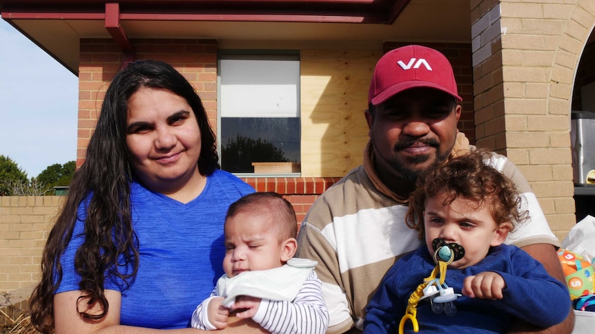 Two young parents sit outside a home with their two babies.