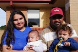 Two young parents sit outside a home with their two babies.