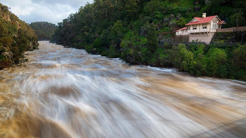 Blurred water speeds through Cataract Gorge, with cottage on the hillside well above the water.