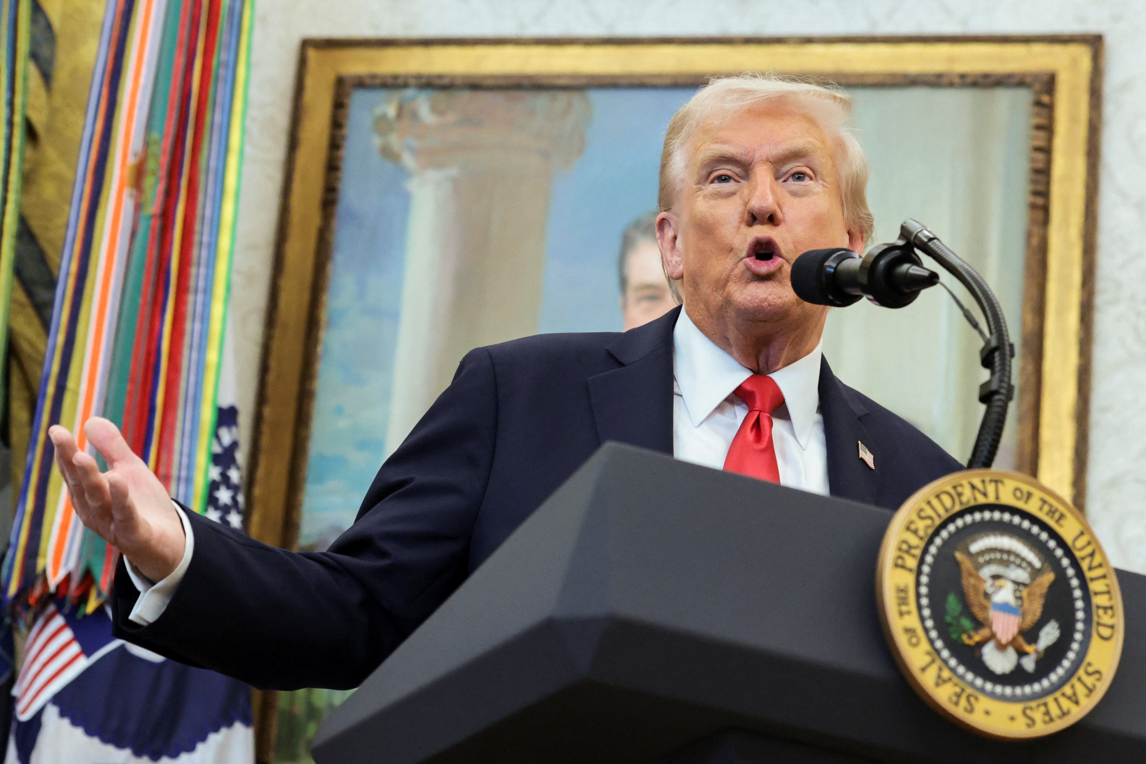 Donald Trump in a dark suit, white shirt and red tie gesturing with his right hand while speaking at a presidential lectern