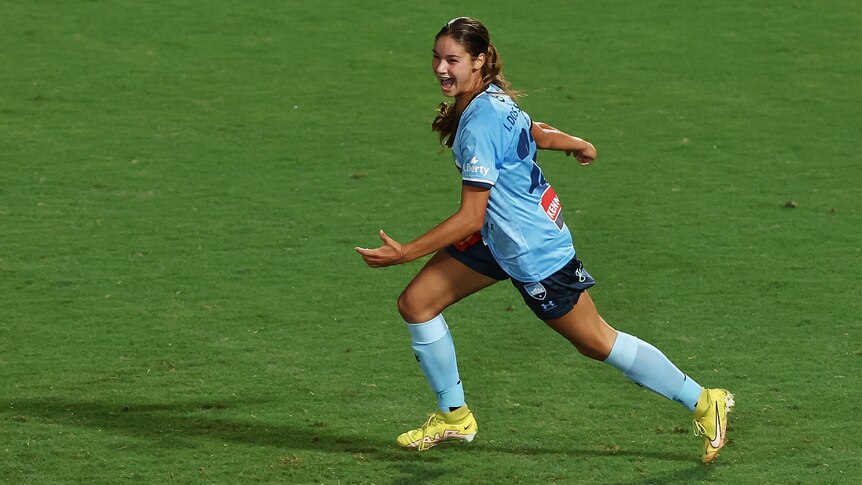 A soccer player wearing light blue runs across some green grass with her arms spread wide in joy