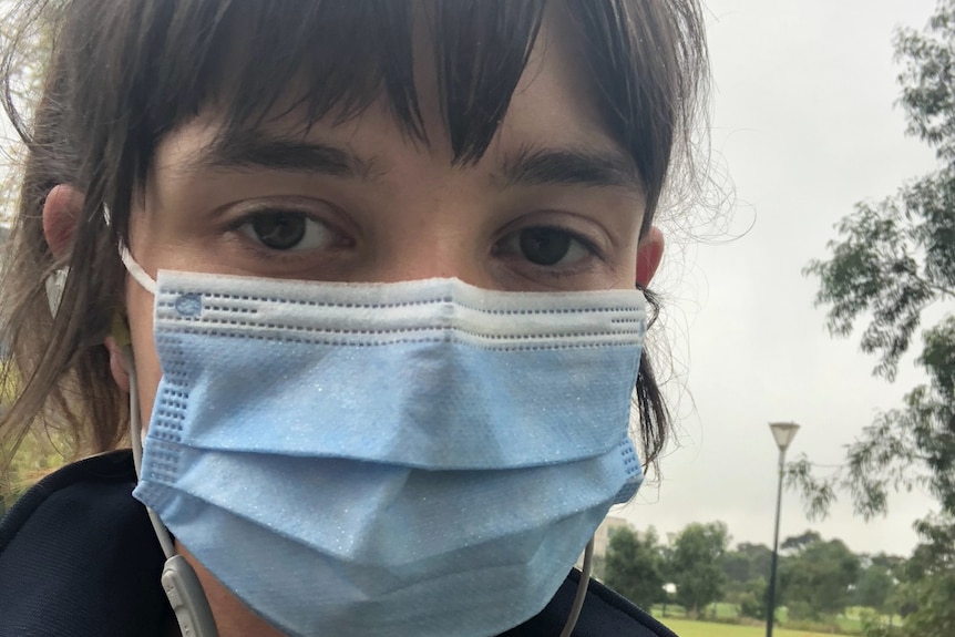 A woman with brown hair appears in a selfie in parkland wearing a blue surgical mask.