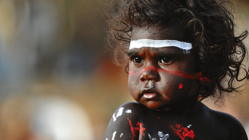 Close-up of Indigenous toddler, smiling, with coloured paint across their face.