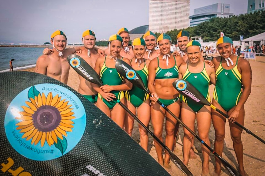 Ten athletes in green and gold swimmers standing on a beach holding ski paddles adorned with sunflower stickers.