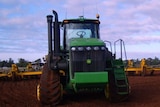 A big green tractor pulling a wide yellow plough through a red dirt paddock