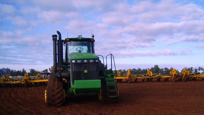 A big green tractor pulling a wide yellow plough through a red dirt paddock