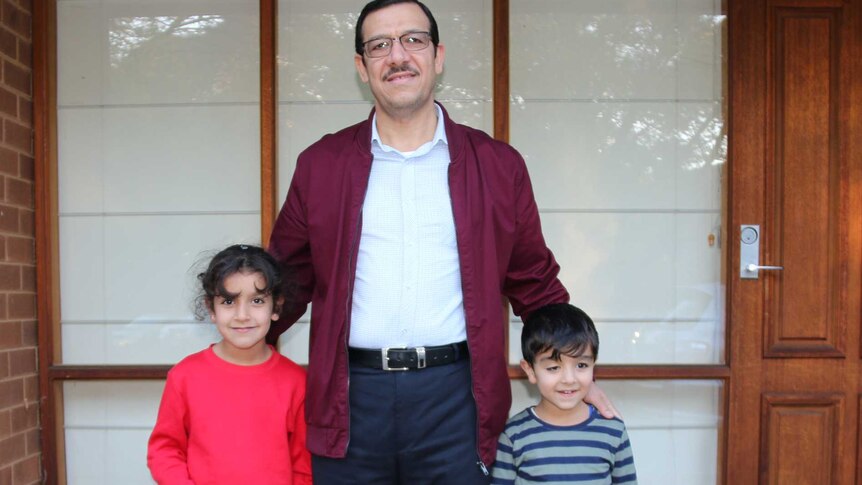 Sameer stands with his two young children out the front of their home.