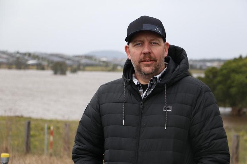 Mid shot of a man wearing a cap and puffer jacket standing in front of floodwaters