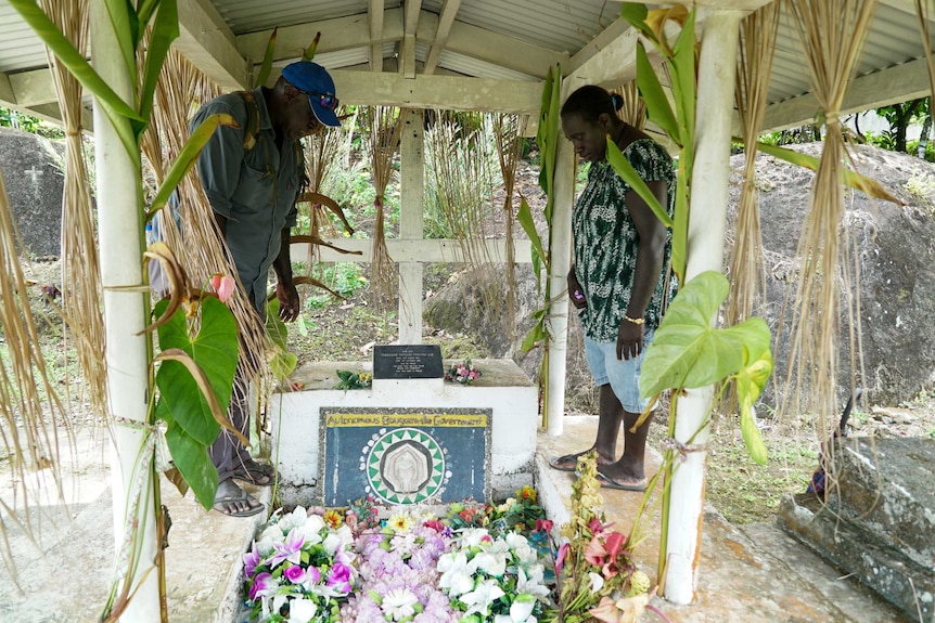 A man and a woman stand at either side of the grave and look down.