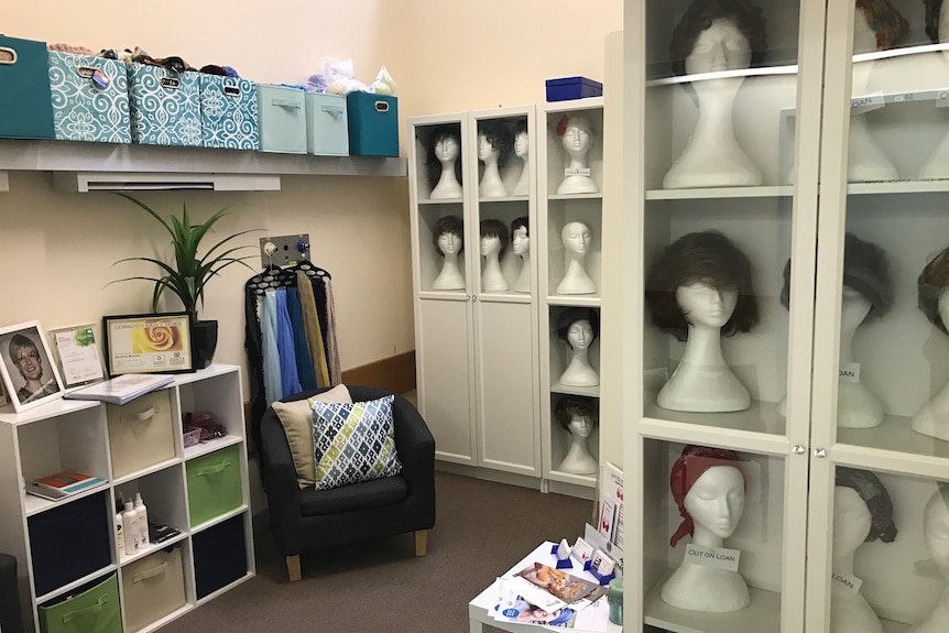 A room with shelves and cupboards full of mannequins wearing different coloured and styled wigs with a chair in the corner