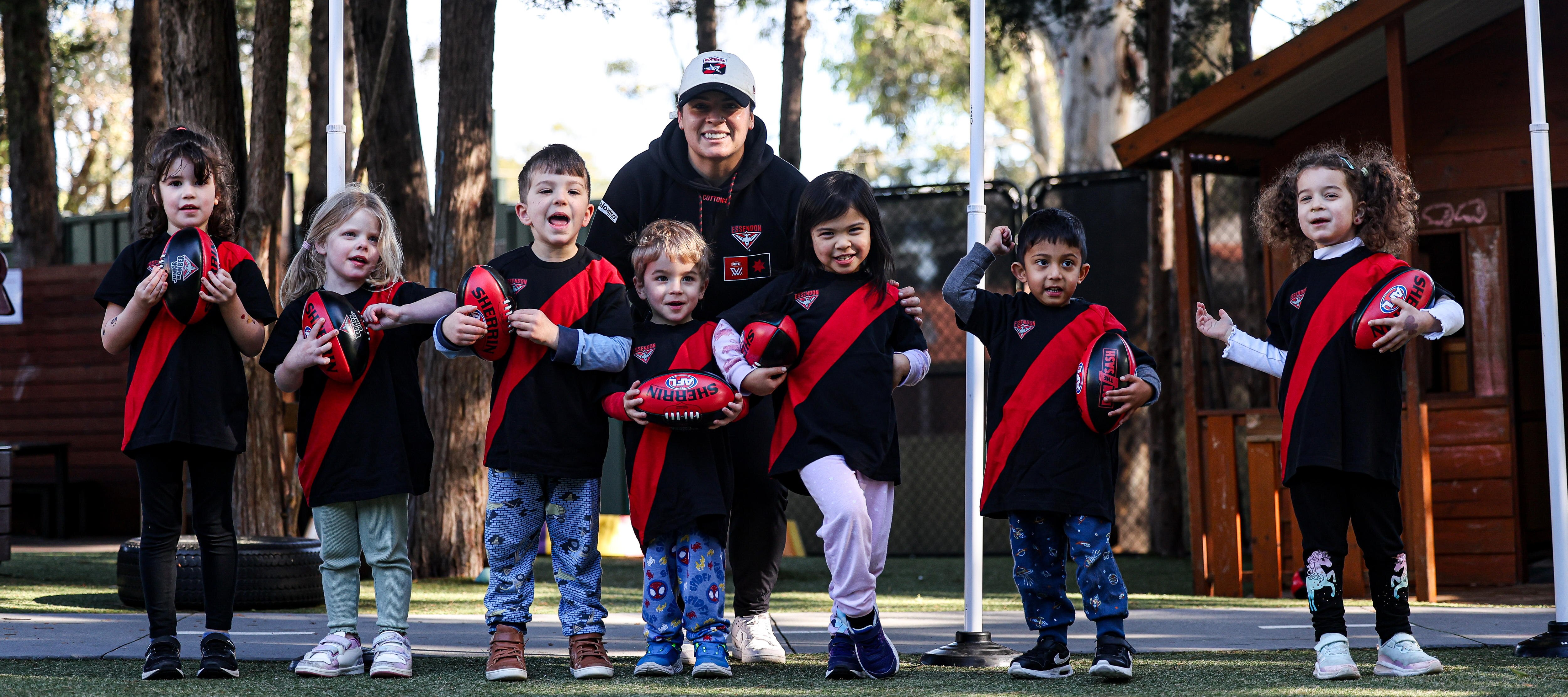 Seven happy young children red and black jerseys stand in a row holding AFL footballs. A woman supervisor stands behind them.