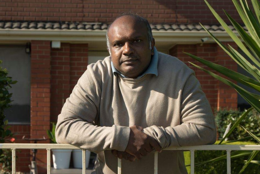 A man leans over a metal fence in his front yard with serious expression on his face