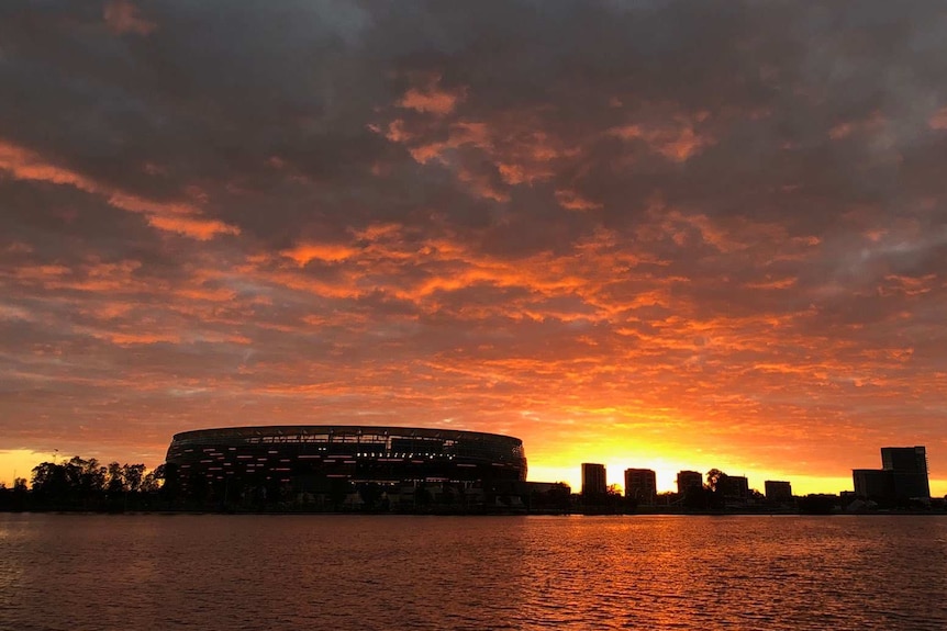 A wide shot showing a glowing red sunrise over Perth Stadium and the surrounding skyline.