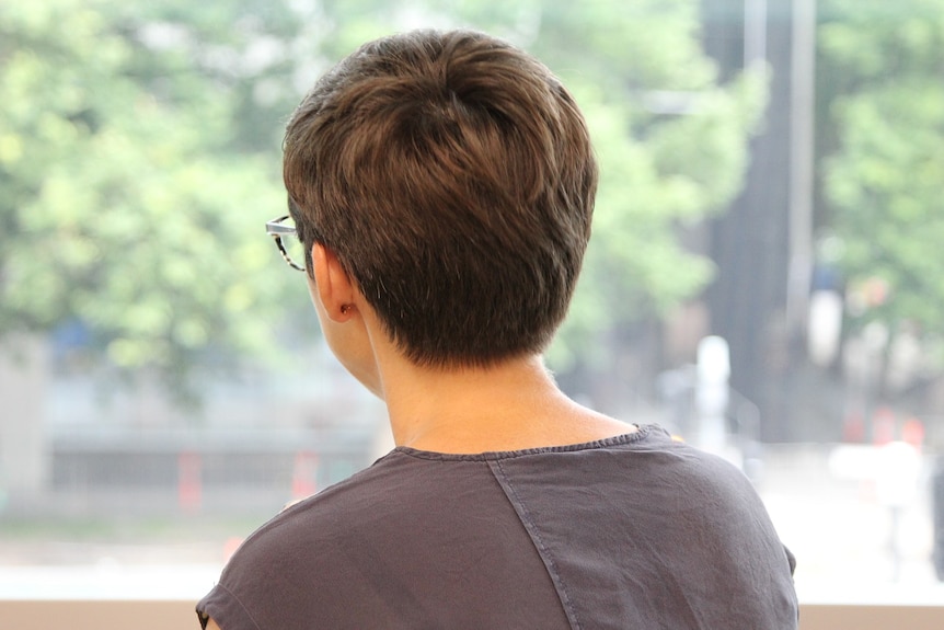 A woman with short hair and glasses faces away from the camera and stares out a window.