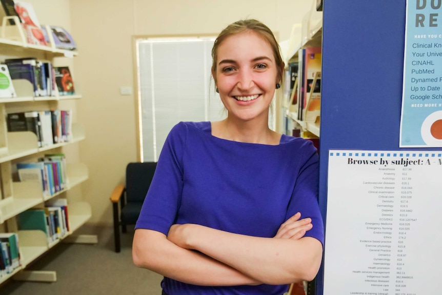 A young fair woman with blonde hair smiles into the camera, she is wearing a royal blue shirt and leans against library shelves.