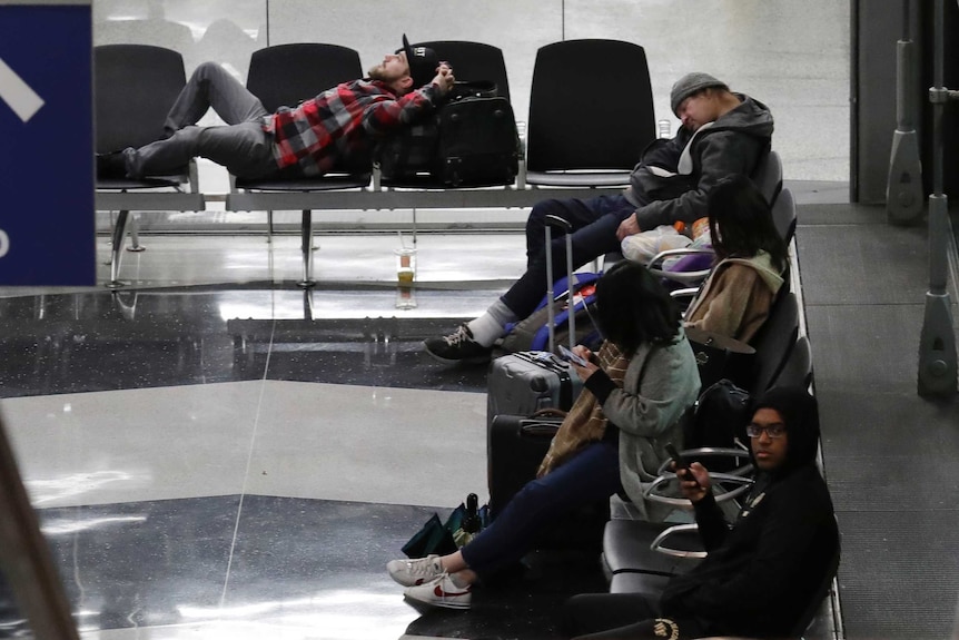 Travellers wait for flights in an airport terminal. Some are sleeping in chairs, others scrolling on mobile phones.