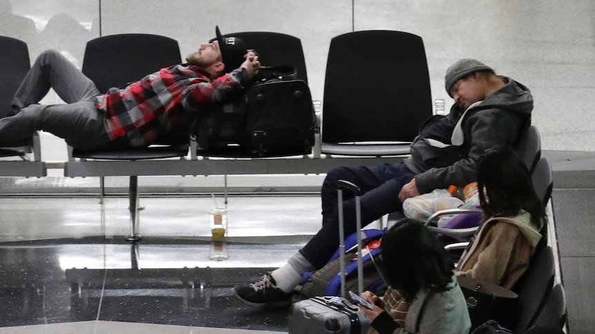 Travellers wait for flights in an airport terminal. Some are sleeping in chairs, others scrolling on mobile phones.