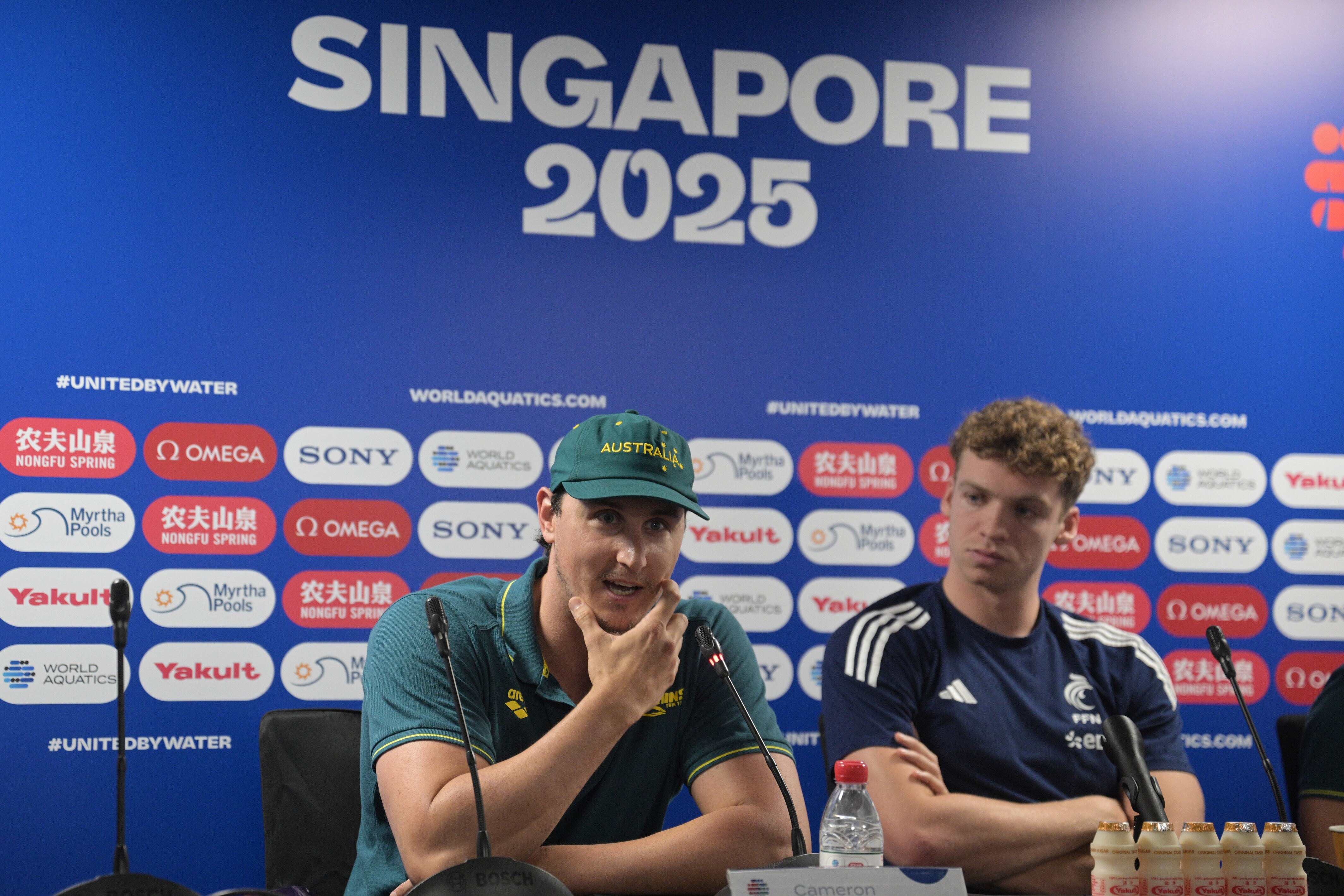 Cameron McEvoy, wears green cap, jersey, speaks at conference with France's Leon Marchand. Singapore 2025 behind.
