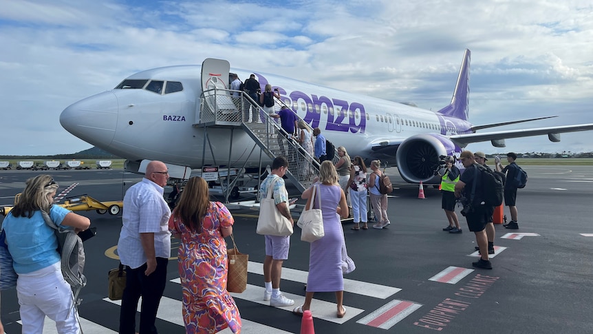 People dressed in summer clothes board plane on tarmac under scattered cloud on a blue sky. Women wear colourful dresses, top.