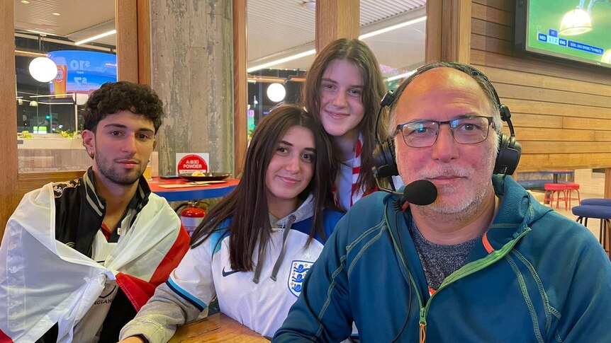 man with spectacles and grey beard at table with two teenaged girls and a boy draped in white flag with red cross