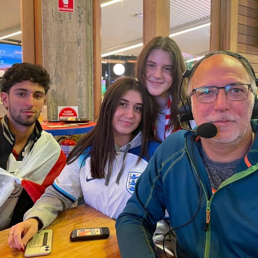 man with spectacles and grey beard at table with two teenaged girls and a boy draped in white flag with red cross