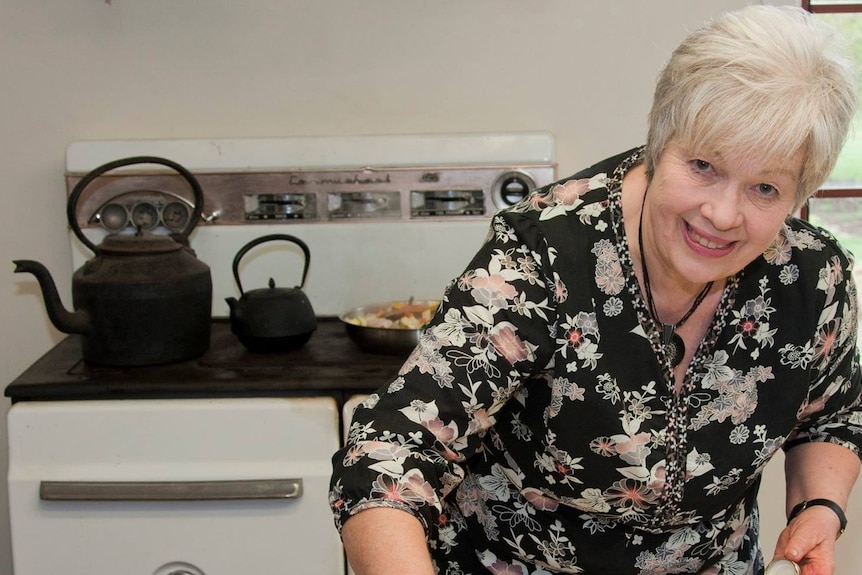 A grey-haired smiling woman in a kitchen leans over various spices and pickles