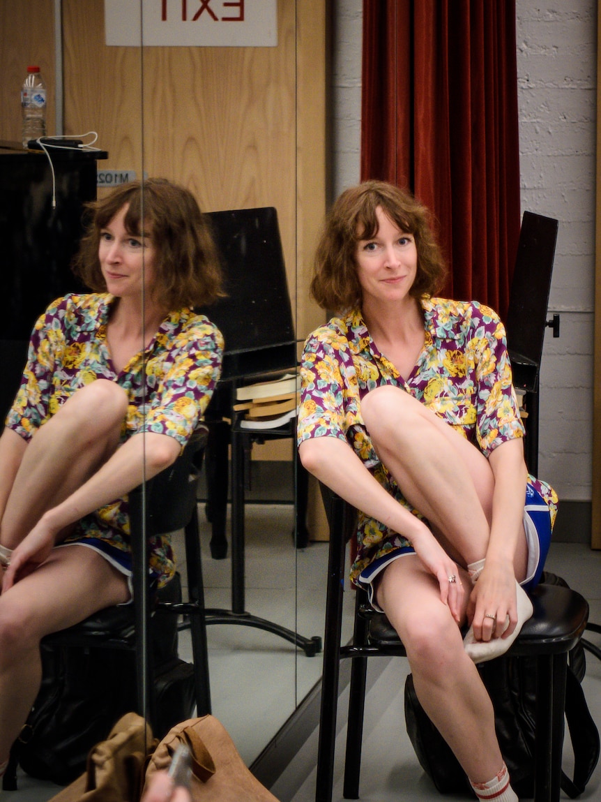 A smiling woman in a bright floral top sits against a chair against a mirror, which reflects her image.