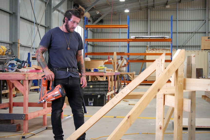 A man in safety googles holds a staple gun as he builds a timber set frame inside a busy warehouse. 