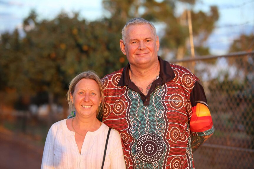A smiling woman with white shirt, blonde hair stands next to a tall man with grey hair wearing an Aboriginal-design shirt.