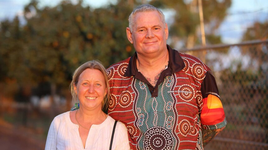A woman with a white shirt, blonde hair stands smiling next to a taller man with grey hair wearing an Aboriginal-patterned shirt
