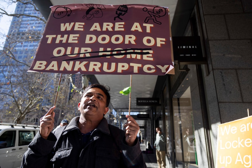 A man holds a poster reading 'WE ARE AT THE DOOR OF (OUR HOME) BANKRUPTCY'.