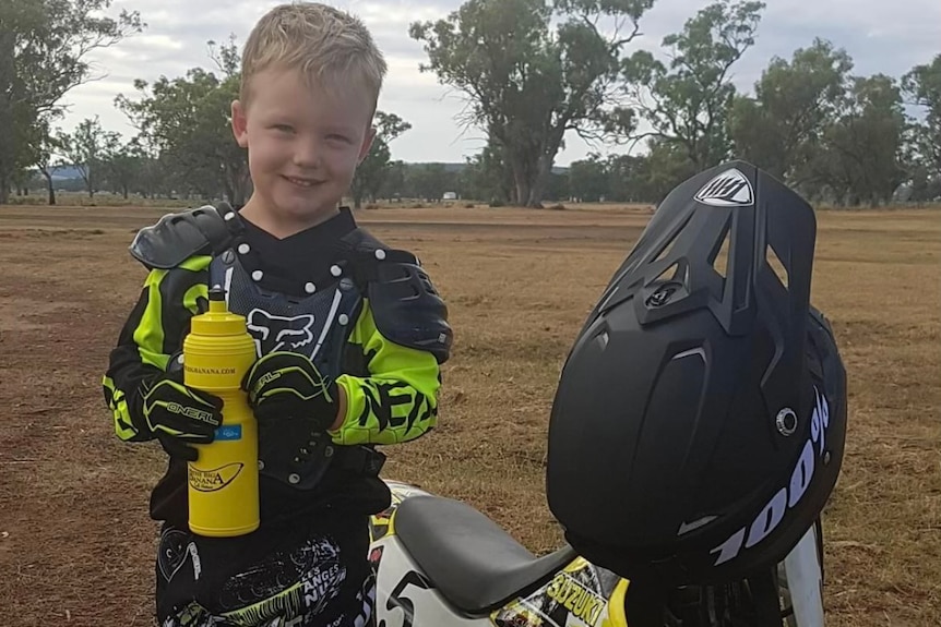A young boy stands next to his motorcycle with a yellow water bottle in his hands