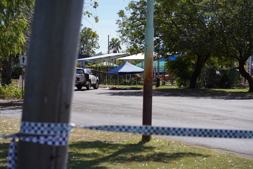 Cordoned police tape around a tree in front of a playground behind a tall metal fence.