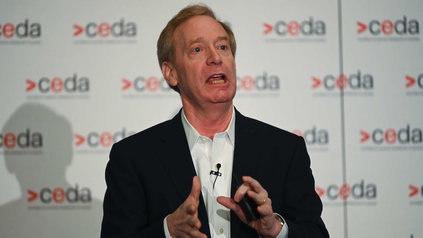 Brad Smith, wearing a black jacket and white shirt, makes a gesture with his hands as he stands speaking.