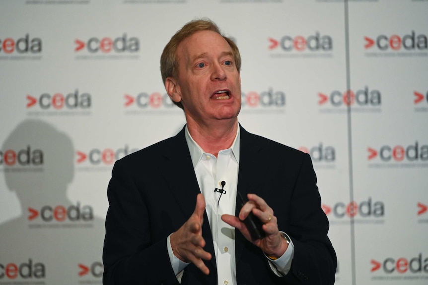 Brad Smith, wearing a black jacket and white shirt, makes a gesture with his hands as he stands speaking.