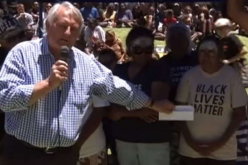 A man is holding a microphone and speaking at a rally during the day with a crowd of people standing beside him