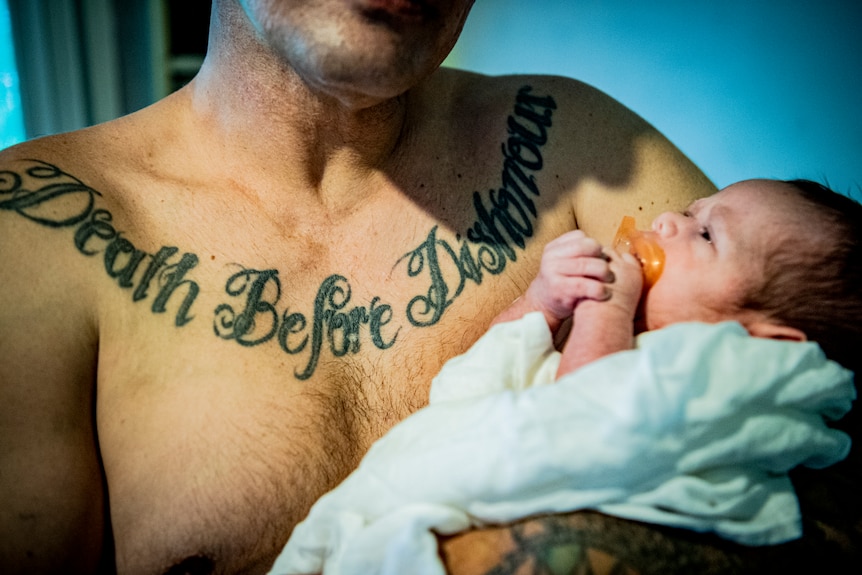 Shirtless man with tattoo across his chest holding a baby.