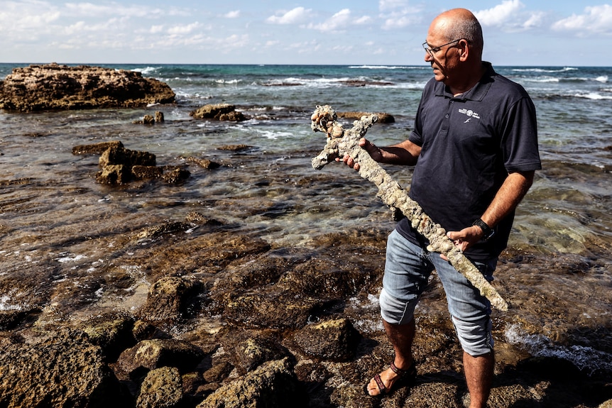 A man standing in the shallows of a rocky outcrop holds a barnacle-encrusted sword.