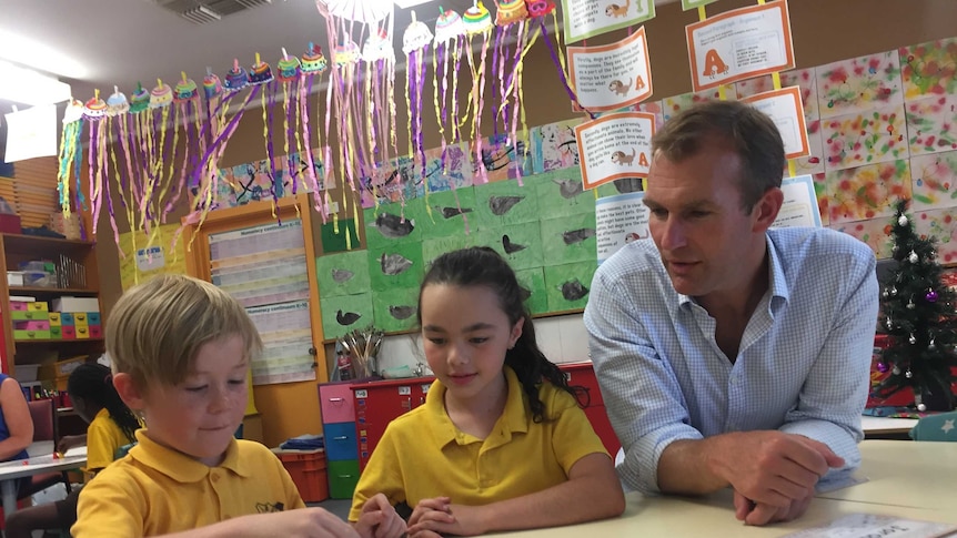 A man in a blue button up shirt sitting at a table with young school students who are working on a maths problem.