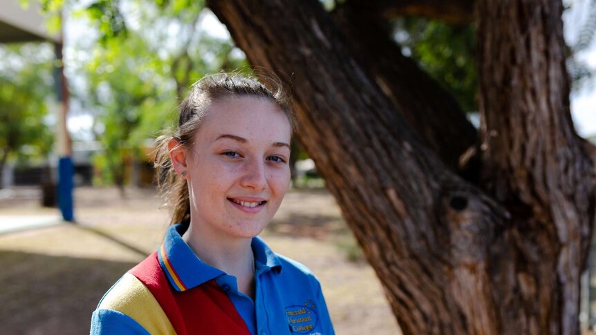 Young school girl, wearing school sports shirt, smiles at camera. Tree and greenery in background.