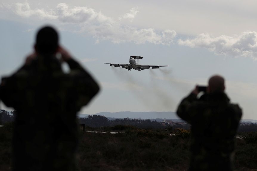 A NATO plane approaches a landing as people in the foreground onlook.