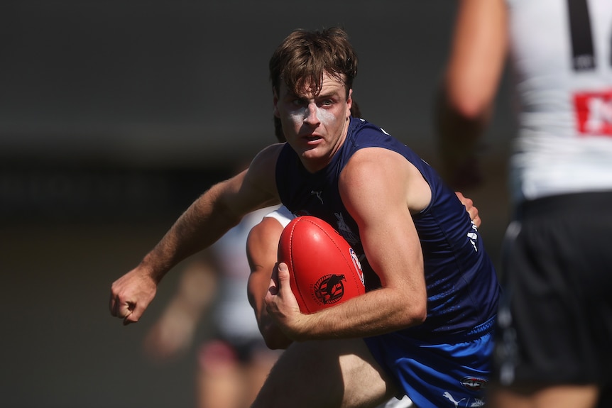 A North Melbourne AFL player runs with the ball tucked in one arm as he clenches his other fist.  