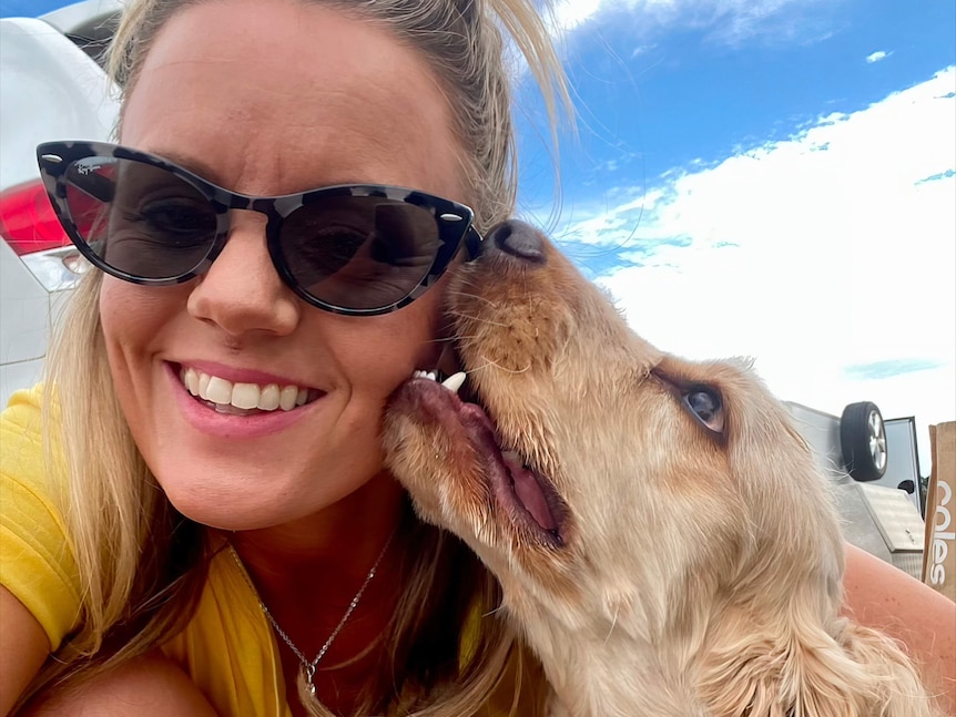 A woman in cat-eye sunglasses poses for a selfie with her dog licking her cheek