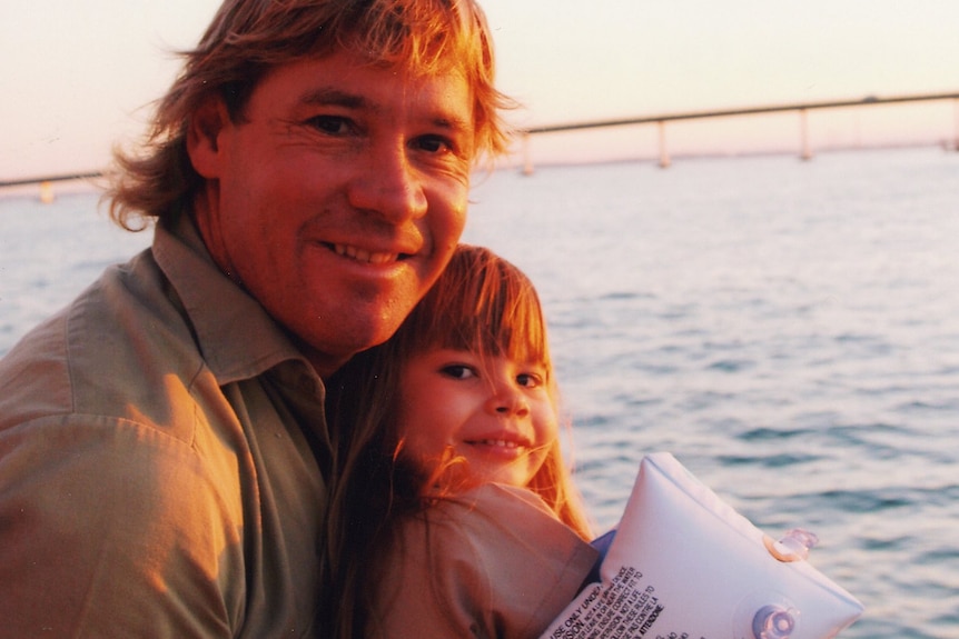 An image of Bindi Irwin wearing floaties as a child being held by her father Steve Irwin with the ocean behind them