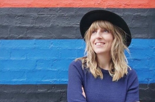 A woman with long blonde hair, black hat and navy jumper stands against coloured painted wall looking up and smiling widely.
