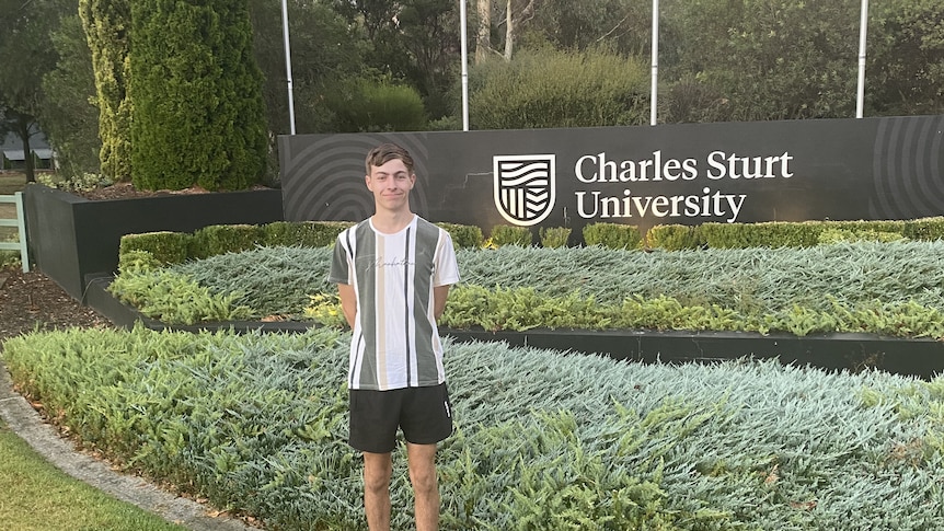 A teenager in a t shirt and shorts standing in front of a garden at a university