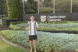 A teenager in a t shirt and shorts standing in front of a garden at a university