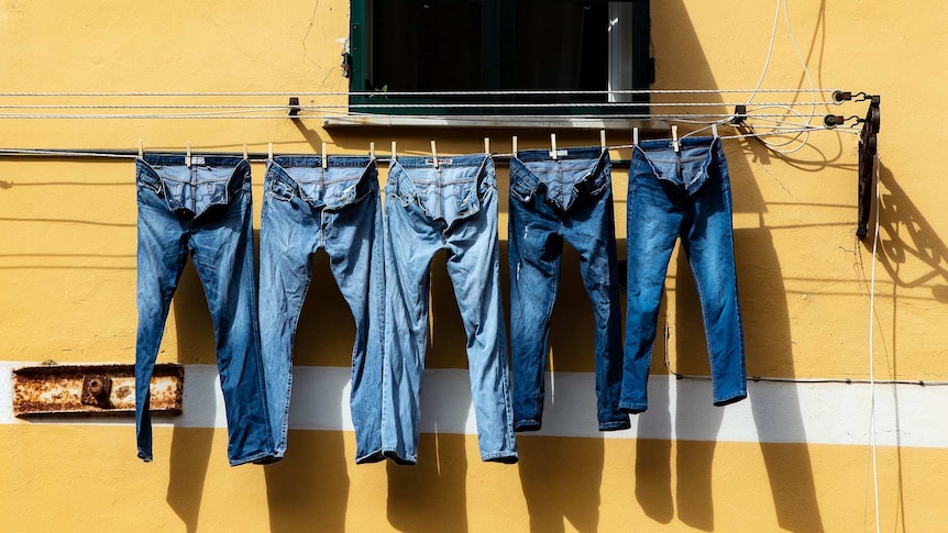 A row of denim jeans hang on the line against a yellow wall to depict theories about denim, such as buying jeans too tight.