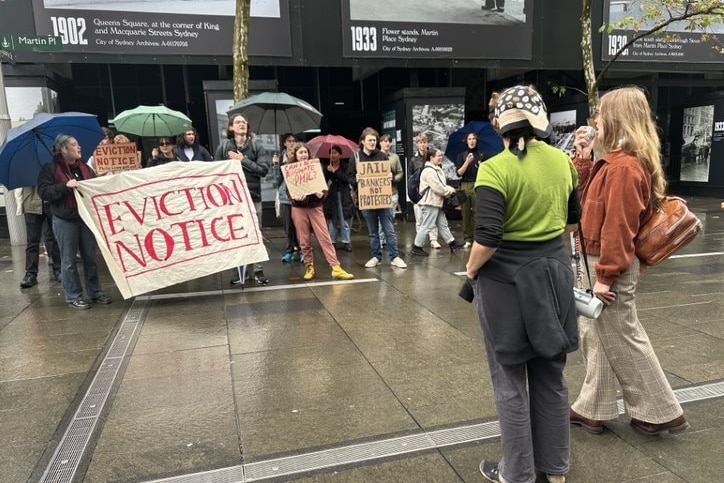 A group of people standing with umbrellas and signs on a wet concrete floor outside a building in the Sydney CBD.